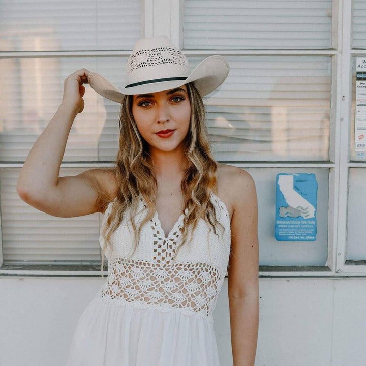 Woman wearing a white lace dress and cowboy hat in front of a building with a sign.